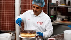 Buddy Valastro carefully decorating a layered cake inside Carlo's Bakery while wearing his chef's uniform and protective hairnet.