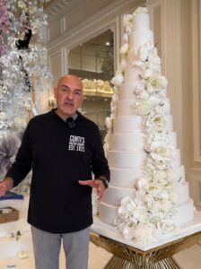 Safet Paljevic standing beside an elegant multi-tiered white wedding cake decorated with cascading flowers at Conti’s Pastry Shoppe.
